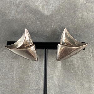 May include: A pair of silver-toned earrings featuring a smooth, curved, triangular shape with a polished metallic finish. The earrings have a dimensional, folded design that tapers to a point on each side, creating a sleek and reflective surface.