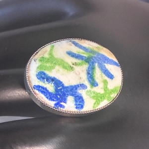 May include: A silver ring with an oval, cream-colored face. The face features a design of blue and green star-like shapes. The ring has a detailed, textured silver band.