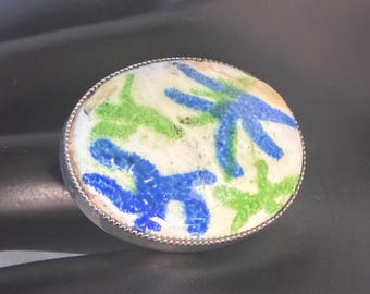 May include: A silver ring with an oval, cream-colored face. The face features a design of blue and green star-like shapes. The ring has a detailed, textured silver band.