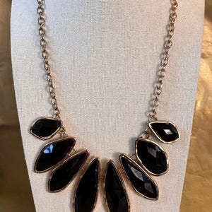May include: A gold-toned necklace with a series of large, black, gem-like pendants. The pendants are set in gold-colored frames and are arranged in a fan shape. The necklace is displayed on a white mannequin.