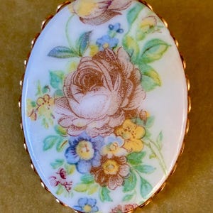May include: Oval brooch with a floral pattern. The design features a central brown rose surrounded by blue, yellow, and pink flowers on a white base. The edge is trimmed with gold-coloured metal.