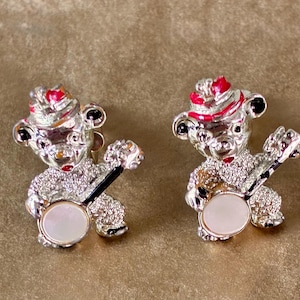 May include: A pair of silver-toned bear-shaped earrings. Each bear wears a red-and-white hat and holds a banjo with a white face. The bears have black accents on their ears and eyes. The earrings are set against a neutral background.