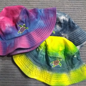 Pode incluir: Três chapéus de balde tie-dye em tons de rosa, azul e amarelo. Cada chapéu apresenta uma aba costurada em branco e um pequeno desenho de tartaruga marinha bordada. Os chapéus são casuais e adequados para proteção solar.