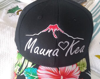 Può includere: Cappellino da baseball nero con un designo ricamato bianco di una montagna con un cuore e il testo "Mauna Kea". Il cappellino ha una stampa floreale sul lato inferiore della visiera.