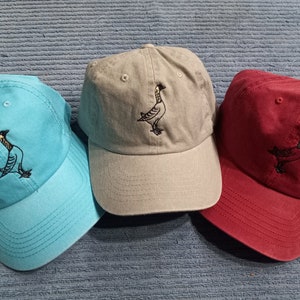 May include: Three baseball caps with a bird design embroidered on the front. The caps are in blue, gray, and red.