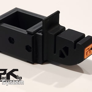 May include: A black plastic 3D printed object with an orange connector. The object has a rectangular base with a small box on top. The object is labeled "PK-clip System".