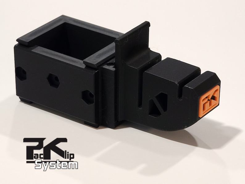 May include: A black plastic 3D printed object with an orange connector. The object has a rectangular base with a small box on top. The object is labeled "PK-clip System".