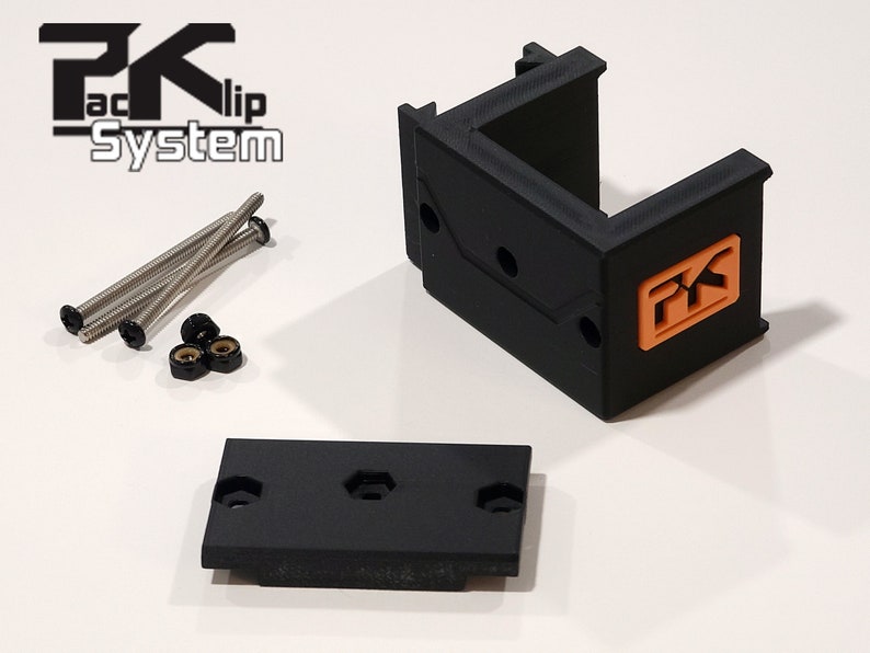 Puede incluir: Un soporte de pl&aacute;stico negro con un logotipo naranja, tornillos y tuercas. El soporte tiene una base rectangular y una parte superior en forma de U. El logotipo est&aacute; en un cuadrado y dice "PK Kli System".