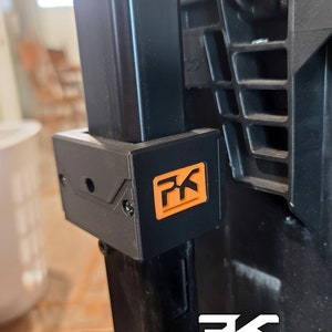 Puede incluir: Primer plano de un soporte de pl&aacute;stico negro con un logotipo naranja "PK". El soporte est&aacute; fijado a un soporte met&aacute;lico negro. Las palabras "PK TacClip System" est&aacute;n impresas en blanco en la parte inferior.