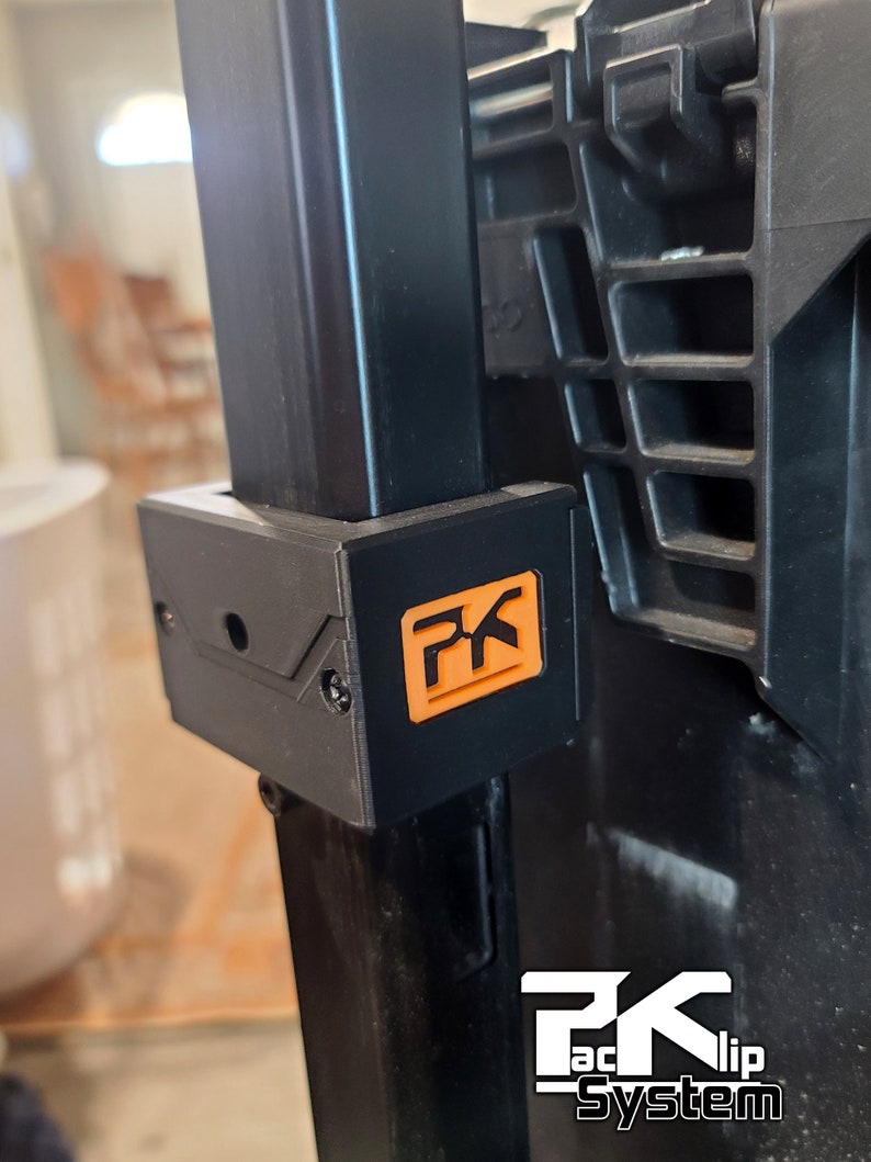 Puede incluir: Primer plano de un soporte de pl&aacute;stico negro con un logotipo naranja "PK". El soporte est&aacute; fijado a un soporte met&aacute;lico negro. Las palabras "PK TacClip System" est&aacute;n impresas en blanco en la parte inferior.