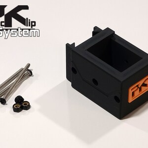 Puede incluir: Un componente de pl&aacute;stico negro con un logotipo naranja, acompa&ntilde;ado de tornillos plateados y tuercas negras. El componente tiene forma rectangular con un interior abierto. El texto "PK Clip System" es visible.