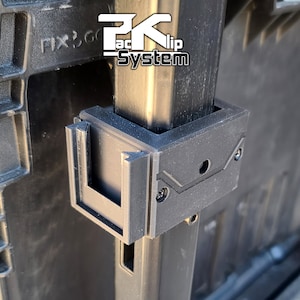 May include: Close-up of a black plastic clip system for attaching items to a metal pole. The clip has a rectangular shape with a slot for the pole and a small hole on the top. The text "Pack-Clip System" is visible on the pole.