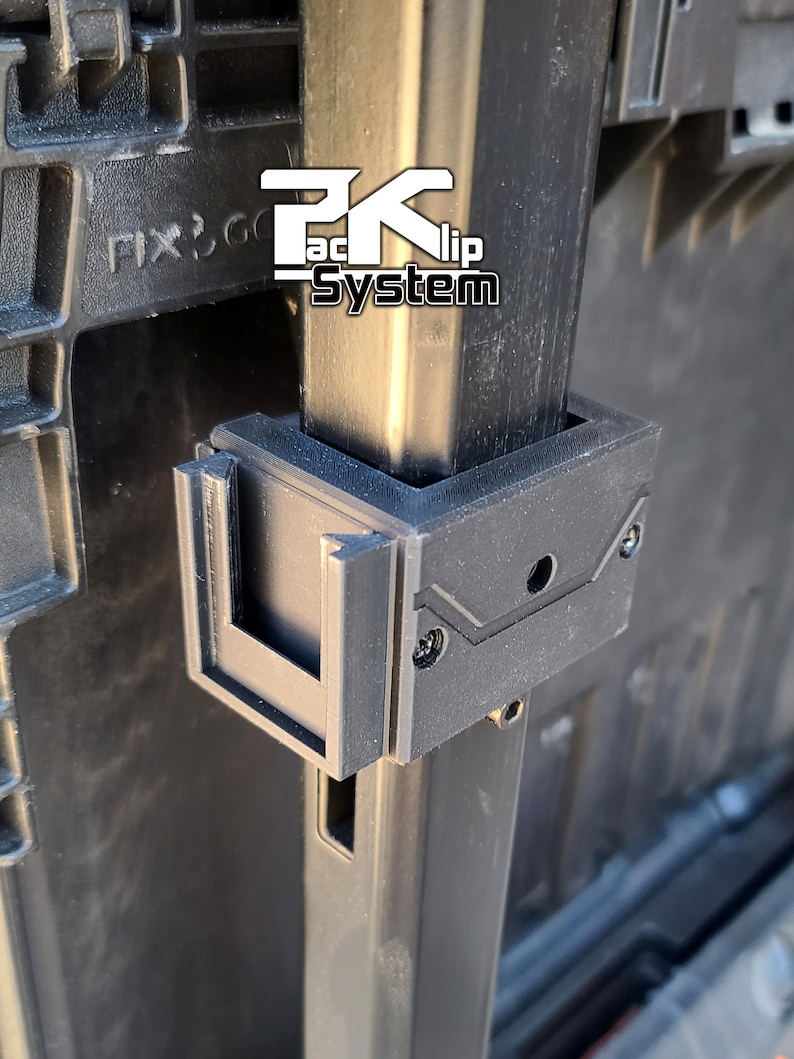 May include: Close-up of a black plastic clip system for attaching items to a metal pole. The clip has a rectangular shape with a slot for the pole and a small hole on the top. The text "Pack-Clip System" is visible on the pole.