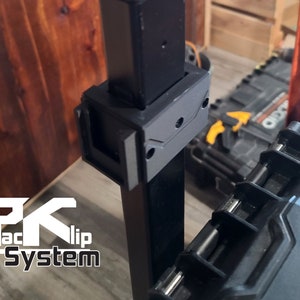 Puede incluir: Sistema de clip Pack negro con el texto "PK Pack Clip System" en blanco. El sistema est&aacute; fijado a un poste de metal negro. Un clip de pl&aacute;stico negro est&aacute; fijado al poste. El fondo incluye una superficie de madera y una caja de herramientas negra.