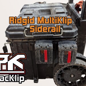 May include: A black Ridgid MultiKlip Siderail toolbox with two black battery packs attached to the side. The toolbox has a black handle and wheels. The toolbox is labelled "PackKlip" in white letters with a red outline.