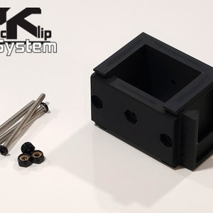 Puede incluir: Soporte de montaje de pl&aacute;stico negro con el texto "PK Clip System". El soporte tiene forma rectangular con interior hueco y varios orificios para tornillos. Incluye tornillos plateados y tuercas negras.