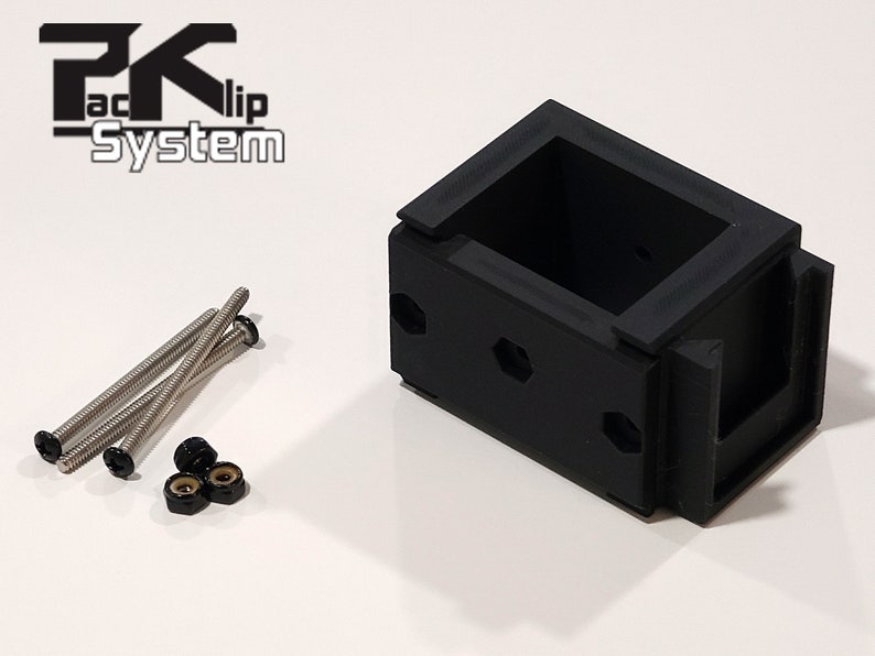 Puede incluir: Soporte de montaje de pl&aacute;stico negro con el texto "PK Clip System". El soporte tiene forma rectangular con interior hueco y varios orificios para tornillos. Incluye tornillos plateados y tuercas negras.