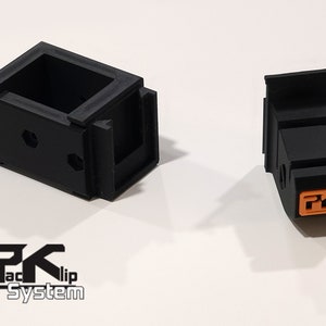 May include: Two black plastic 3D printed parts, one with a rectangular shape and the other with a more angular shape. The rectangular part has a small opening on one side. The angular part has an orange logo with the letters "PK" on it.