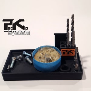 May include: A black plastic organizer with a blue container of solder, a small orange and black holder with the letters "PK" on it, and a variety of drill bits and screws. The organizer has the text "PKaclip System" on the top.
