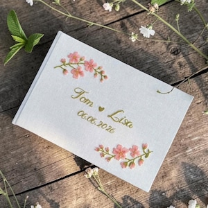 Op de afbeelding: Een witte huwelijksuitnodiging met geborduurde roze bloemaccenten. De tekst "Tom ♥ Lisa" en de datum "06.06.2026" zijn in gouden letters geschreven. De uitnodiging ligt op een rustiek houten oppervlak, omgeven door groen en witte bloemen.