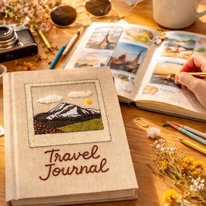 May include: A beige linen-covered travel journal featuring an embroidered square patch of a mountain landscape with white clouds and a yellow sun. The words Travel Journal are embroidered in brown thread below the patch on the front cover.