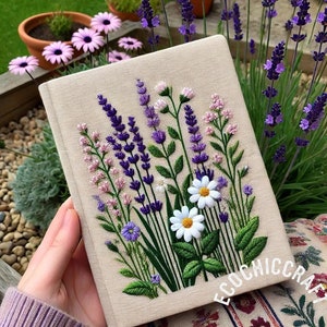 May include: A beige linen notebook featuring a hand-stitched embroidery design of lavender flowers, daisies, and greenery. The notebook is resting on a surface with a floral pattern.
