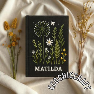 Könnte beinhalten: Ein schwarzer Buchumschlag mit gestickten Blumenmustern, darunter ein Löwenzahn, weiße Blumen und grüne Blätter. Der Titel "MATILDA" ist in Weiß dargestellt, mit "ECOCHIC CRAFT" in einem kreisförmigen Design unten.