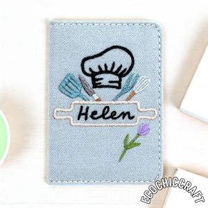 May include: A pale blue embroidered notebook cover with a chef's hat, rolling pin, and kitchen tools. The name "Helen" is embroidered below the rolling pin. A purple tulip is embroidered on the bottom. The cover is made of a textured fabric.