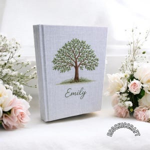 以下が含まれることがあります： 刺繍された木の模様が施された、明るい灰色のハードカバーの日記。木には緑の葉と茶色の幹があり、下に「Emily」という名前が刺繍されています。日記は白とピンクの花に囲まれています。