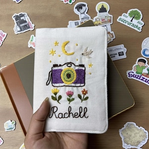 Puede incluir: Una funda blanca para pasaporte con un dise&ntilde;o de c&aacute;mara bordada en morado y amarillo, con una luna creciente, estrellas y flores. El nombre "Rachell" est&aacute; bordado debajo. La funda est&aacute; sujeta con una mano.