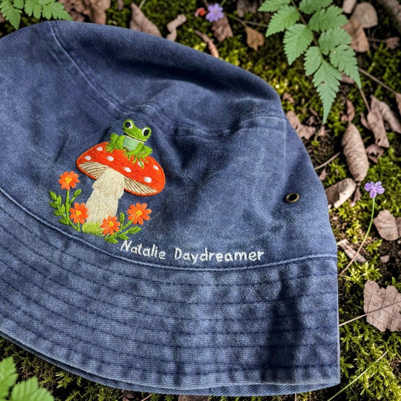 May include: A navy blue bucket hat featuring an embroidered design of a green frog sitting on a red and white mushroom surrounded by orange flowers. The text "Natalie Daydreamer" is embroidered in white thread below the mushroom design.