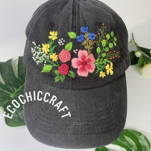 May include: A dark gray baseball cap with embroidered flowers in pink, yellow, blue, and white. The brim of the cap features the text "ECOCHIC CRAFT" in white. The floral embroidery adds a colorful, handcrafted touch to the hat.