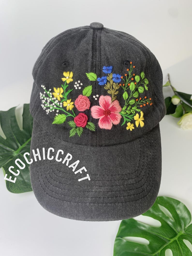 May include: A dark gray baseball cap with embroidered flowers in pink, yellow, blue, and white. The brim of the cap features the text "ECOCHIC CRAFT" in white. The floral embroidery adds a colorful, handcrafted touch to the hat.