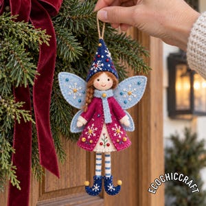 Può includere: Un ornamento a forma di fata in feltro con un cappello a punta blu scuro ricamato con fiocchi di neve bianchi, ali azzurre, un cappotto magenta e gambe a righe. La figura ha capelli castani e indossa scarpe blu arricciate.