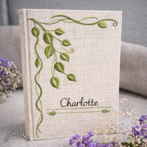 May include: A linen-covered book with green embroidered leaves and vines. The name "Charlotte" is embroidered in black script. The book is standing upright, with a floral arrangement in the background.