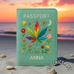 May include: A teal passport holder with the word "PASSPORT" embroidered in white. The cover features a colourful embroidered cannabis leaf and floral designs. The name "ANNA" is embroidered below the leaf. The background is a beach scene.