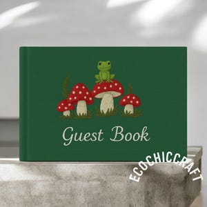 Può includere: Un libro degli ospiti verde con un disegno ricamato di funghi rossi con pois bianchi e una rana verde. Le parole "Guest Book" sono scritte in corsivo bianco.