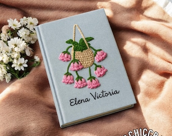 Könnte beinhalten: Hellblaues Notizbuch mit gesticktem Blumendesign. Der Einband zeigt einen geflochtenen Korb mit rosa Blüten und grünen Blättern. Der Name "Elena Victoria" ist in schwarzer Schrift geschrieben. Das Notizbuch liegt auf einer pfirsichfarbenen Decke.
