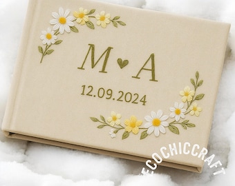 May include: A beige wedding guest book with embroidered floral details. The cover displays the initials "M ❤️ A" and the date "12.09.2024" in gold. White and yellow daisies with green leaves decorate the corners of the book.