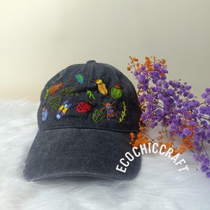 Op de afbeelding: Een zwarte denim baseballcap met kleurrijke geborduurde insecten, waaronder lieveheersbeestjes, kevers en vlinders. De cap heeft een gebogen klep en een wit geborduurd logo met de tekst "Ecochiccraft".