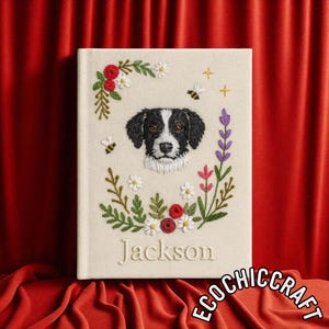 May include: Cream-colored journal with an embroidered portrait of a black and white dog, surrounded by floral and bee accents. The name "Jackson" is embroidered below the dog portrait. The journal is set against a red backdrop.