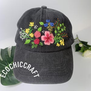 May include: A dark gray baseball cap with embroidered flowers in pink, red, yellow, and blue. The text "ECOCHIC CRAFT" is printed in white on the brim. The cap has a curved brim and a button on top.