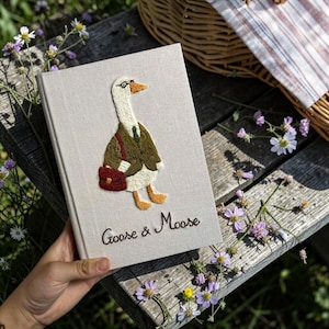 Puede incluir: Un libro de tapa dura color beige claro con una oca blanca bordada que lleva una chaqueta verde, un bolso rojo oscuro y unas gafas peque&ntilde;as. El texto "Goose & Moose" est&aacute; bordado en hilo marr&oacute;n oscuro debajo de la oca en la cubierta de tela.