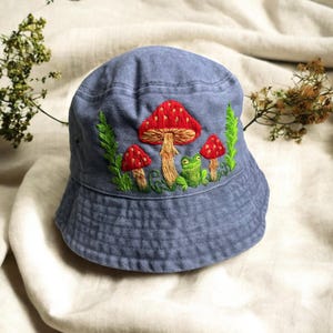 May include: A blue bucket hat featuring detailed embroidery of three red mushrooms with white spots and a small green frog. The design includes green foliage accents surrounding the mushrooms and the frog, creating a nature-inspired motif on the front.