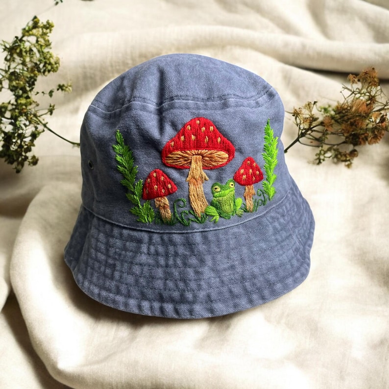 May include: A blue bucket hat featuring detailed embroidery of three red mushrooms with white spots and a small green frog. The design includes green foliage accents surrounding the mushrooms and the frog, creating a nature-inspired motif on the front.