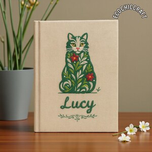 Könnte beinhalten: Ein beiges Notizbuch mit einem gestickten Design einer grünen Katze mit floralen Details und roten Blumen. Der Name "Lucy" ist unter der Katze gestickt. Das Notizbuch hat den Text "ECOCHIC CRAFT" oben rechts.