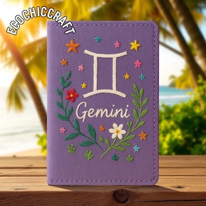 May include: A purple Gemini passport holder with embroidered floral and star accents. The design includes the Gemini symbol and the word "Gemini" in white thread. The holder is on a wooden surface with a blurred beach background.