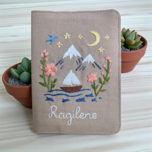 Puede incluir: Funda para pasaporte beige con dise&ntilde;o bordado. Presenta un velero sobre el agua, monta&ntilde;as, estrellas y una luna creciente. Tambi&eacute;n est&aacute;n bordadas flores rosas y el nombre "Ragilene". Dos peque&ntilde;as suculentas en macetas a cada lado.
