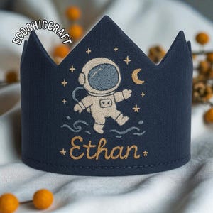 以下が含まれることがあります： 宇宙飛行士、星、三日月が刺繍されたネイビーブルーの王冠。名前「Ethan」は金色の糸で刺繍されています。子供向けの気まぐれなアクセサリーです。