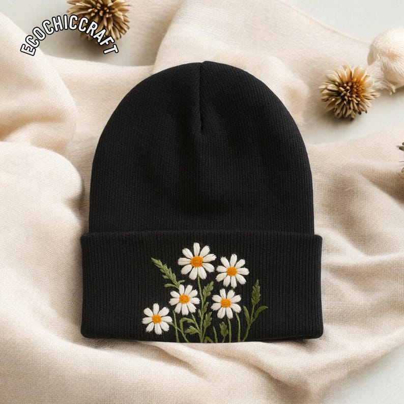 May include: A black beanie with a folded brim, embroidered with a cluster of white daisies with yellow centers and green stems and leaves. The text "ECOCHIC CRAFT" is printed on the top left of the image.